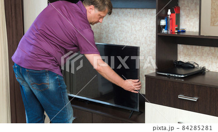 A tall blond man in jeans and a lilac shirt installs a new large LCD TV on the bedside table A tall blond man in jeans and a lilac shirt installs a new large LCD TV on the bedside table 84285281
