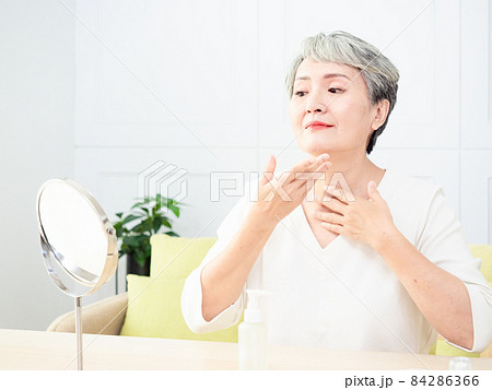 Happy 50s middle aged asian woman touching face skin looking in mirror reflection. Smiling mature old lady pampering, healthy moisturized skin care, aging beauty, skincare treatment cosmetics concept. 84286366