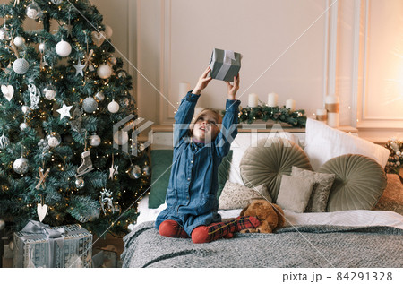 a little beautiful girl is sitting on the bed next to the Christmas tree holding a gift high up 84291328