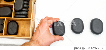Hot stones for massage in a wooden box next to stone in the hand of masseur isolated on white background top view. Hot stones for massage in a wooden box next to stone in the hand of masseur isolated on white background top view. 84292257