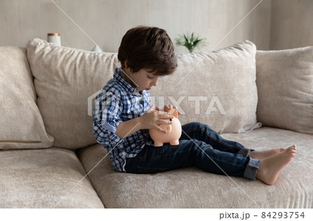 Preschool boy sit on sofa throw coins into piggy bank Preschool boy sit on sofa throw coins into piggy bank 84293754