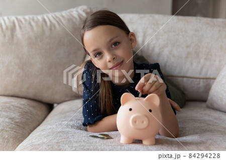 Cute thrifty little girl put coin into piggy bank Cute thrifty little girl put coin into piggy bank 84294228