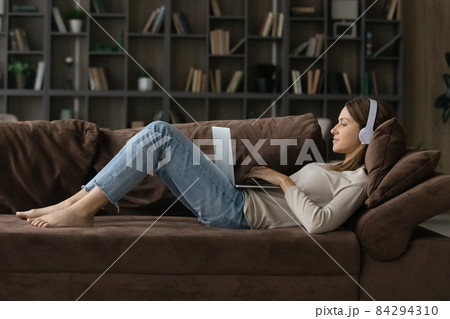 Woman lying on comfy sofa wear headphones using laptop 84294310