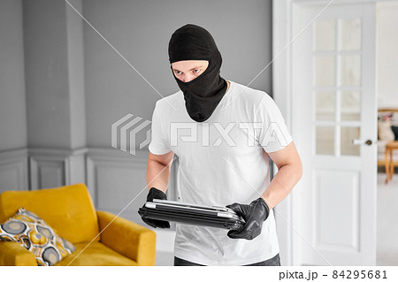 Thief with black balaclava stealing modern expensive laptop from apartment. Masked face. Man burglar stealing tv set from house Thief with black balaclava stealing modern expensive laptop from apartment. Masked face. Man burglar stealing tv set from house 84295681