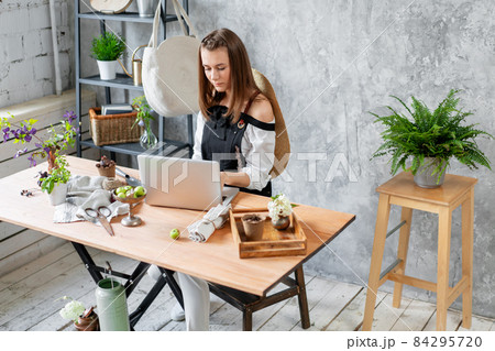 Young female freelancer works on a laptop, responds to messages in social networks. Flower delivery concept. Professional worker in a flower shop. 84295720