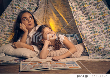 Mother and daughter are sitting in a teepee tent with some pillows and reading stories. Happy family. Mother and daughter are sitting in a teepee tent with some pillows and reading stories. Happy family. 84307792