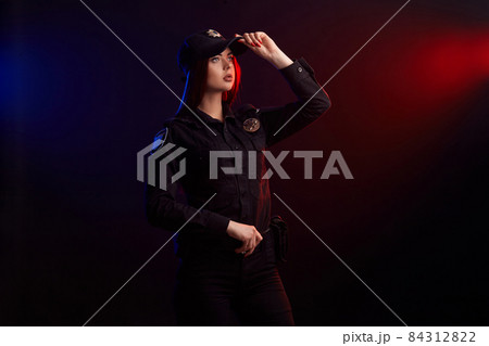 Serious female police officer is posing for the camera against a black background with red and blue backlighting. Serious female police officer is posing for the camera against a black background with red and blue backlighting. 84312822