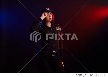 Serious female police officer is posing for the camera against a black background with red and blue backlighting. Serious female police officer is posing for the camera against a black background with red and blue backlighting. 84312823