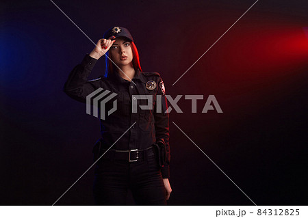 Serious female police officer is posing for the camera against a black background with red and blue backlighting. Serious female police officer is posing for the camera against a black background with red and blue backlighting. 84312825