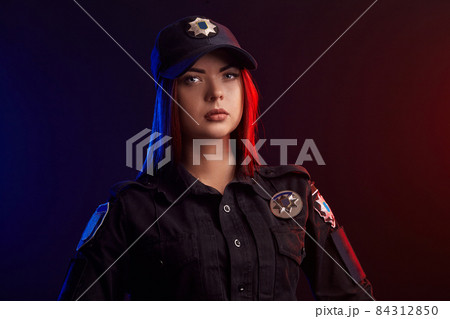 Close-up shot of serious female police officer posing for the camera against a black background with red and blue backlighting. Close-up shot of serious female police officer posing for the camera against a black background with red and blue backlighting. 84312850