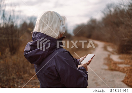 Middle age beautiful blonde woman listening to music using headphones in the city autumn park. 84315442