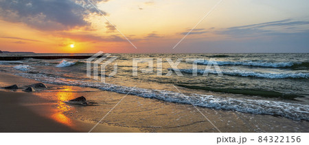 Sunset in the wavy sea with a wooden breakwater and stones on the sandy shore, beautifully colored sky 84322156