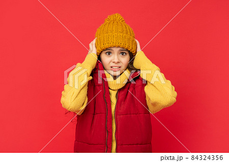 kid in knitted winter hat and down vest feel cold on red background, fashion 84324356