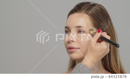 Makeup artist or stylist applies foundation or concealer to the face of the female model. Visagiste  applying cosmetic foundation base on a face. 84325876