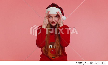 Woman in sweater Santa Christmas hat fooling around having closing eyes with hand and spying through Woman in sweater Santa Christmas hat fooling around having closing eyes with hand and spying through 84326819