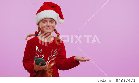 Girl toddler in Christmas Santa hat with mobile phone showing pointing empty place, advertising area Girl toddler in Christmas Santa hat with mobile phone showing pointing empty place, advertising area 84327471