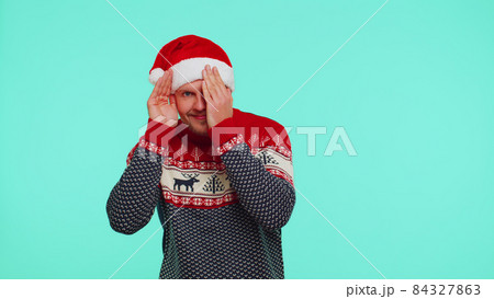 Man in sweater Santa Christmas hat fooling around having closing eyes with hand and spying through Man in sweater Santa Christmas hat fooling around having closing eyes with hand and spying through 84327863