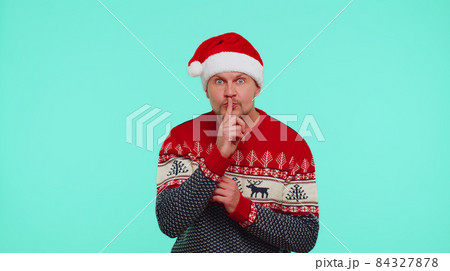 Man wears red Christmas sweater presses index finger to lips makes silence gesture sign, secret 84327878