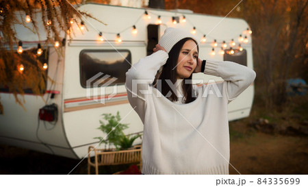 A pensive Caucasian woman in a white knitted sweater and a hat is resting near a motorhome in a warm autumn. Travel by camper. 84335699
