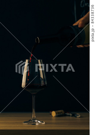 Wine is pouring into a wine glass from a bottle on a dark background, the cork and corkscrew lie side by side. Front view. 84338736