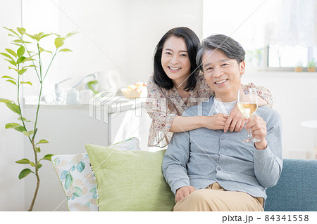 リビングでソファに座ってワインを持つミドルエイジの男性と後ろからバックハグをする女性のポートレート 84341558