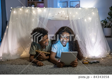 Cheerful African American brother and sister lying on carpet behind table covered with fabric and lights and using tablet 84343058