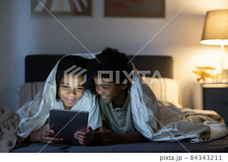 Positive black siblings lying under duvet on bed and using tablet while browsing internet at night Positive black siblings lying under duvet on bed and using tablet while browsing internet at night 84343211