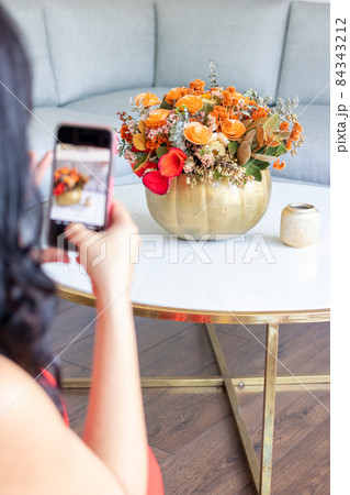 Brunette woman is taking a photo of a cute floral bouquet inside a cabbage-painted pumpkin. 84343212