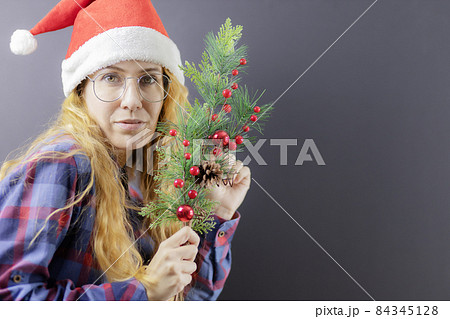 A blonde lady with glasses and a Santa hat holds an elegant spruce branch. Christmas poster A blonde lady with glasses and a Santa hat holds an elegant spruce branch. Christmas poster 84345128