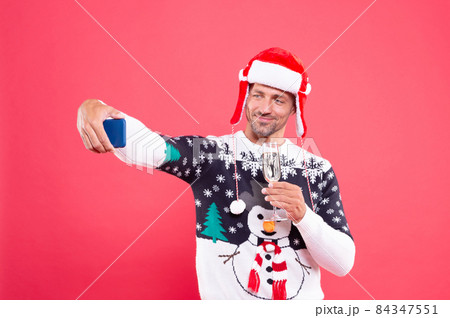 cheerful guy in winter sweater and hat making selfie on phone with champagne on red background, xmas cheerful guy in winter sweater and hat making selfie on phone with champagne on red background, xmas 84347551