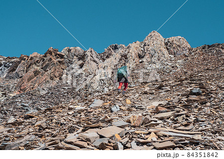 Solo trekking in the mountains. Man with a large backpack is climbing heavily on loose rock trail. Adventure solo traveling lifestyle concept, active weekend vacations on the wild nature. 84351842