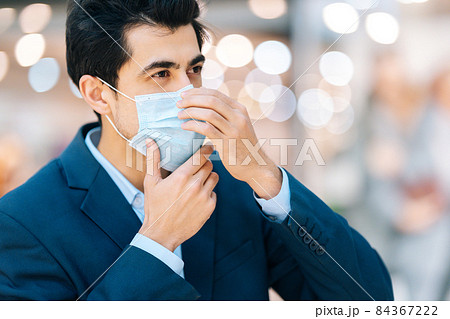 Close-up side view of confident young man in stylish suit wearing protection face mask looking away in hall of mall centre, blurred background. Close-up side view of confident young man in stylish suit wearing protection face mask looking away in hall of mall centre, blurred background. 84367222