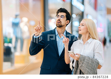 Beautiful young couple searching store in shopping mall using mobile phone, looking around and pointing fingers. 84367228