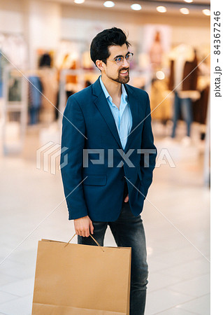 Vertical portrait of smiling elegance bearded young man in stylish suit and glasses holding shopping paper bags with purchases and looking away. 84367246