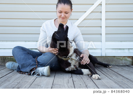 Picture of seated brunette woman in jeans and white jacket stroking black dog on white background wooden wall 84368040