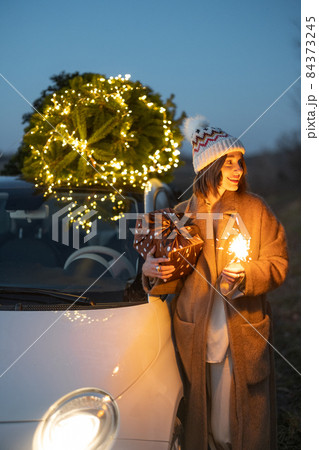 Woman with sprklers near car and Christmas tree outdoors Woman with sprklers near car and Christmas tree outdoors 84373245