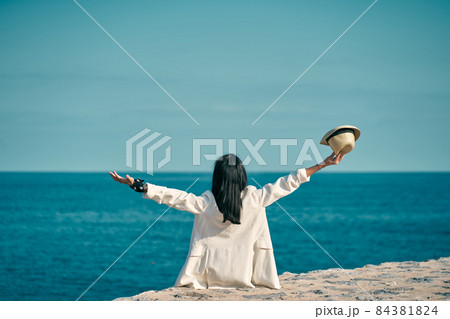 young woman sitting looking out to sea with arms up and hat in hand young woman sitting looking out to sea with arms up and hat in hand 84381824
