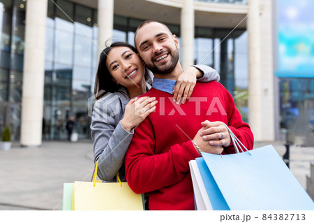 Affectionate interracial couple with shopper bags smiling and hugging near big mall, outdoors Affectionate interracial couple with shopper bags smiling and hugging near big mall, outdoors 84382713