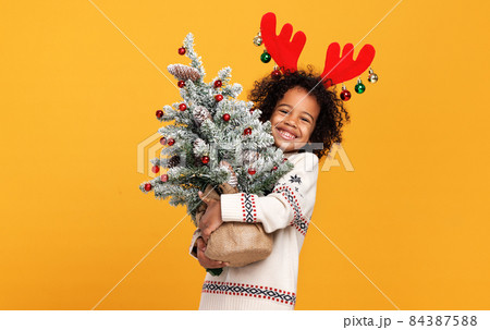 Little preschool african american kid boy in red deer antlers holding snowy Xmas tree 84387588