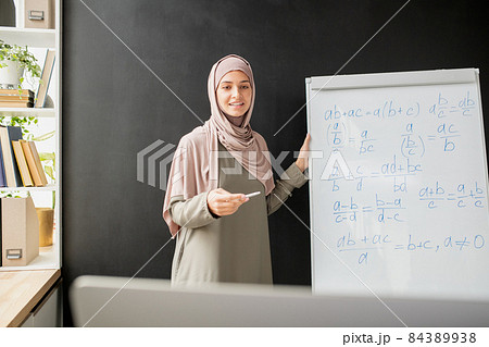 Happy young teacher in hijab standing by whiteboard with algebraic formula and equations Happy young teacher in hijab standing by whiteboard with algebraic formula and equations 84389938