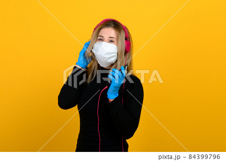Portrait of a girl in a medical mask, which puts on a rubber glove. Yellow and grey background. 84399796