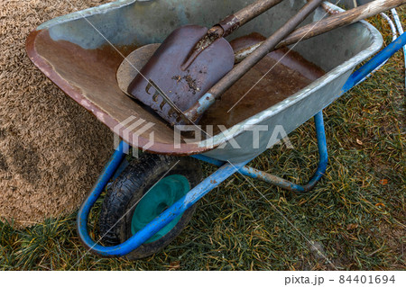 A utility wheelbarrow with tools for construction and gardening. Concept: building material, garden and vegetable garden, own farm, village and farm, agriculture. A utility wheelbarrow with tools for construction and gardening. Concept: building material, garden and vegetable garden, own farm, village and farm, agriculture. 84401694