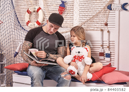 Handsome tattoed man is spending time with his little cute daughter. Reading fairytales while daughter is sitting nearby. Happy family. 84403582