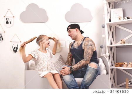 Funny time Tattoed father in a cap and his child are playing at home. Dad is doing his daughter's hair in her bedroom. Family holiday and togetherness Funny time Tattoed father in a cap and his child are playing at home. Dad is doing his daughter's hair in her bedroom. Family holiday and togetherness 84403630