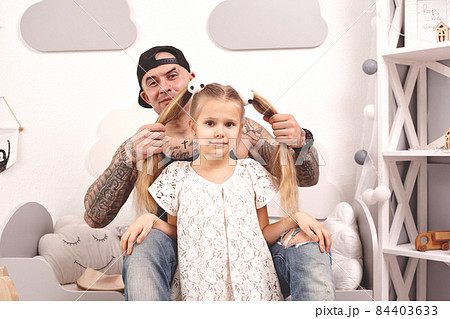 Funny time Tattoed father in a cap and his child are playing at home. Dad is doing his daughter's hair in her bedroom. Family holiday and togetherness 84403633