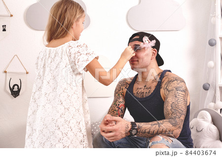 Funny time Tattoed father in a cap and his child are playing at home. Cute girl is doing makeup to her dad in her bedroom. Family holiday togetherness Funny time Tattoed father in a cap and his child are playing at home. Cute girl is doing makeup to her dad in her bedroom. Family holiday togetherness 84403674