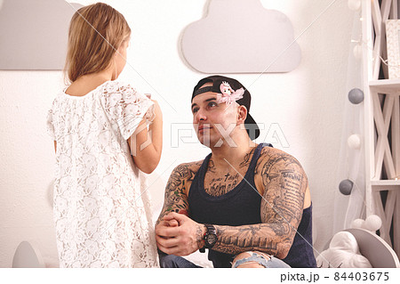 Funny time Tattoed father in a cap and his child are playing at home. Cute girl is doing makeup to her dad in her bedroom. Family holiday togetherness Funny time Tattoed father in a cap and his child are playing at home. Cute girl is doing makeup to her dad in her bedroom. Family holiday togetherness 84403675