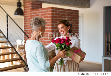 Happy adult daughter bringing gift and bouquet to senior mother indoors at home. Happy adult daughter bringing gift and bouquet to senior mother indoors at home. 84405117
