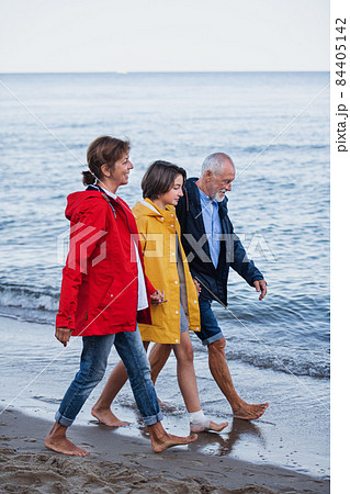 Side view of senior couple holding hands with their preteen granddaughter and walking on sandy beach 84405142