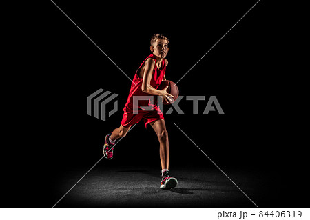 Full-length portrait of teen boy, basketball player in motion, training, running with ball isolated over black background. Goal scoring 84406319